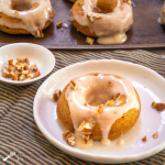 Top down photo of a pumpkin donut with cream cheese glaze and garnished with pecans on a white plate over a striped green napkin. A baking sheet of more donuts sit in the background.