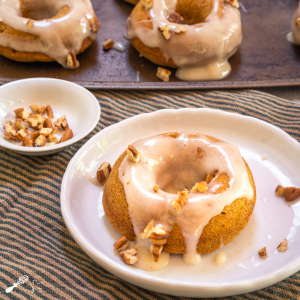 Top down photo of a pumpkin donut with cream cheese glaze and garnished with pecans on a white plate over a striped green napkin. A baking sheet of more donuts sit in the background.