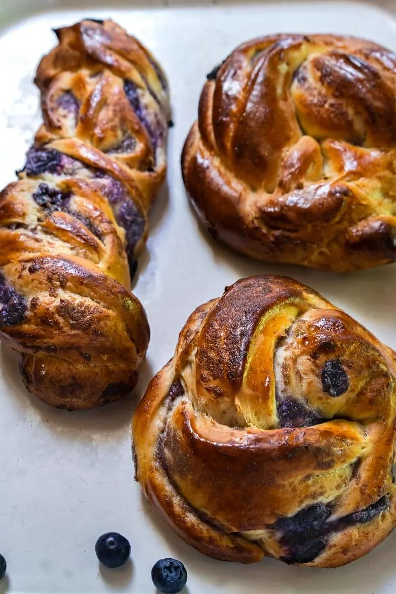 Two round blueberry twist rolls and one twist long twist roll sitting on a white tray. Scattered blueberries are around around the rolls.