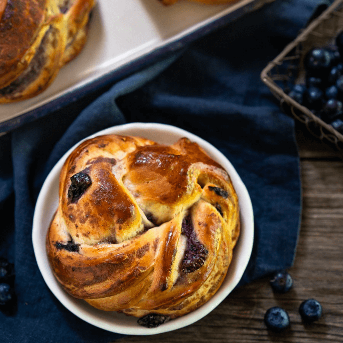 Top down view of a blueberry twist roll sitting on a white plate over a blue napkin over a wooden background. Blueberries are scattered around the plate with more rolls in the background.