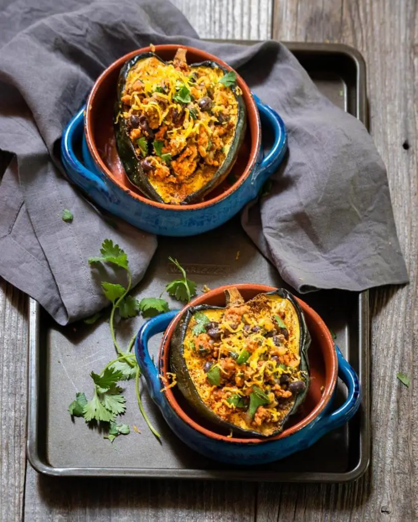 Top-down view of two blue bowls containing a half acorn squash stuffed with chicken chili sitting on a baking sheet over a wooden background. A gray napkin is in the background and fresh cilantro is scattered about the bowls.