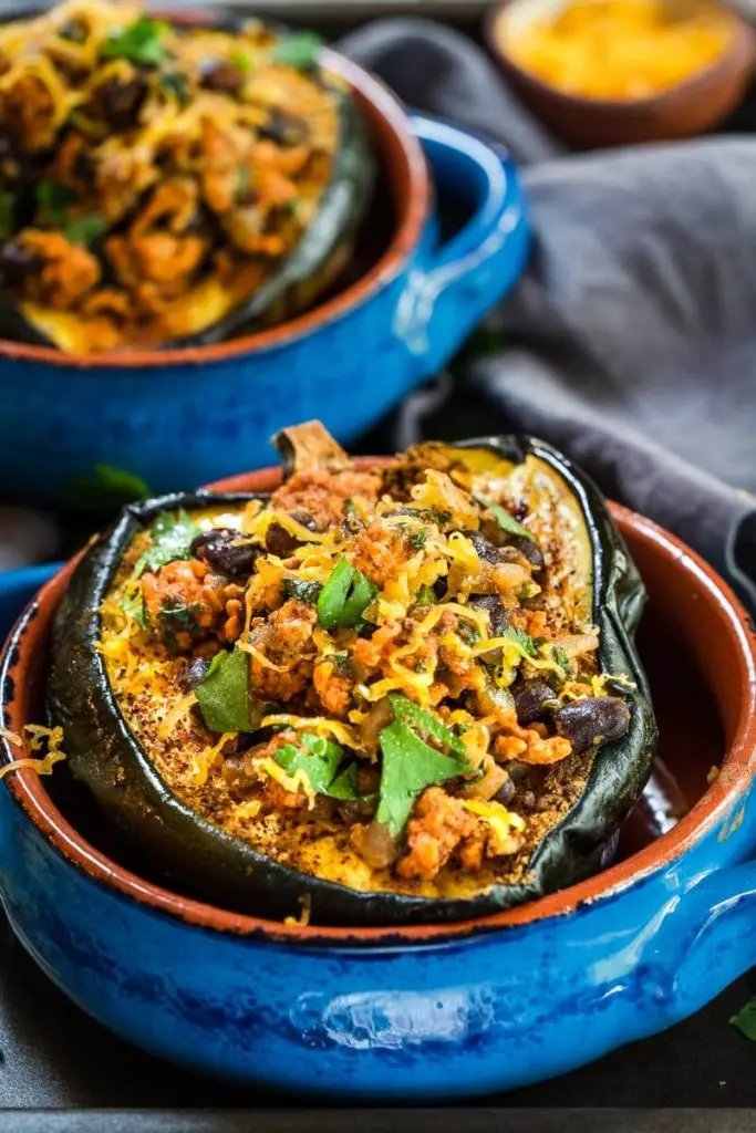 Close-up front view of a baked half of an acorn squash brushed with chili powder and stuffed with chicken chili and garnished with cheese and cilantro sitting in a blue bowl with an adobe red edge. A second baked squash is in the background next to a bowl of shredded cheese sitting over a gray napkin.