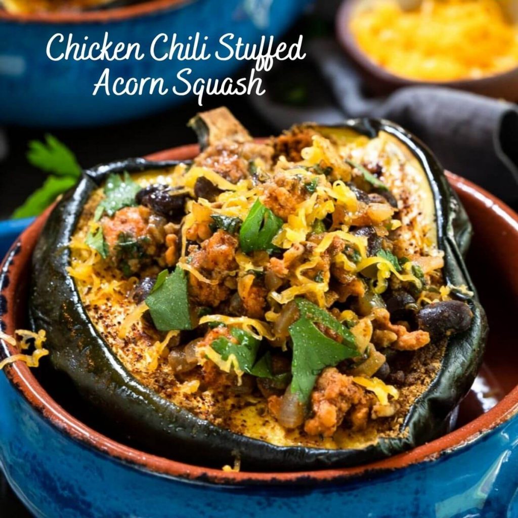 A close up shot of a baked half of an acorn squash brushed with chili powder and stuffed with chicken chili then garnished with cheese and cilantro sitting in a blue bowl with an adobe red edge. The title "Chicken Chili Stuffed Acorn Squash" is in the upper left corner.