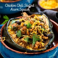 A close up shot of a baked half of an acorn squash brushed with chili powder and stuffed with chicken chili then garnished with cheese and cilantro sitting in a blue bowl with an adobe red edge. The title 