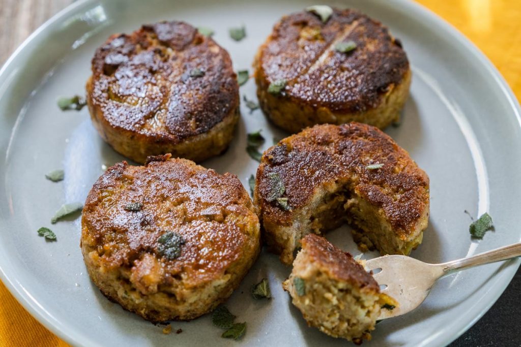 4 Fried Dressing Patties garnished with fresh sage on a gray plate. One Pattie has a fork-filled bite out of it.