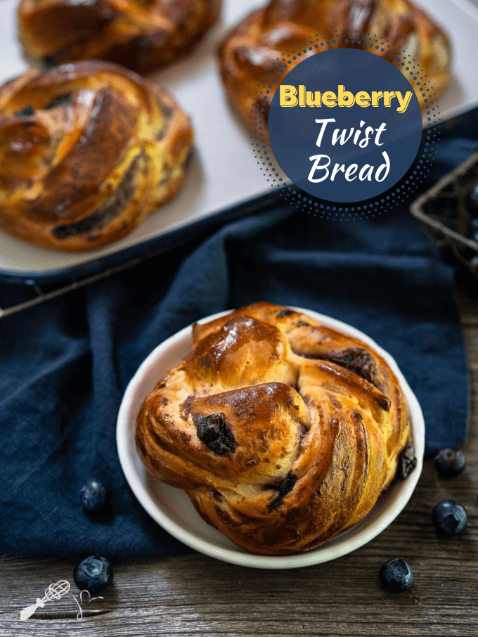 Top down view of a blueberry twist roll sitting on a white plate over a blue napkin over a wooden background. Blueberries are scattered around the plate with more rolls in the background. The title "Blueberry Twist Bread" is in the upper right corner.