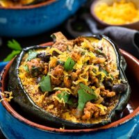A baked half of an acorn squash brushed with chili powder and stuffed with chicken chili and garnished with cheese and cilantro sitting in a blue bowl with an adobe red edge. A second baked squash is in the background next to a bowl of shredded cheese sitting over a gray napkin.