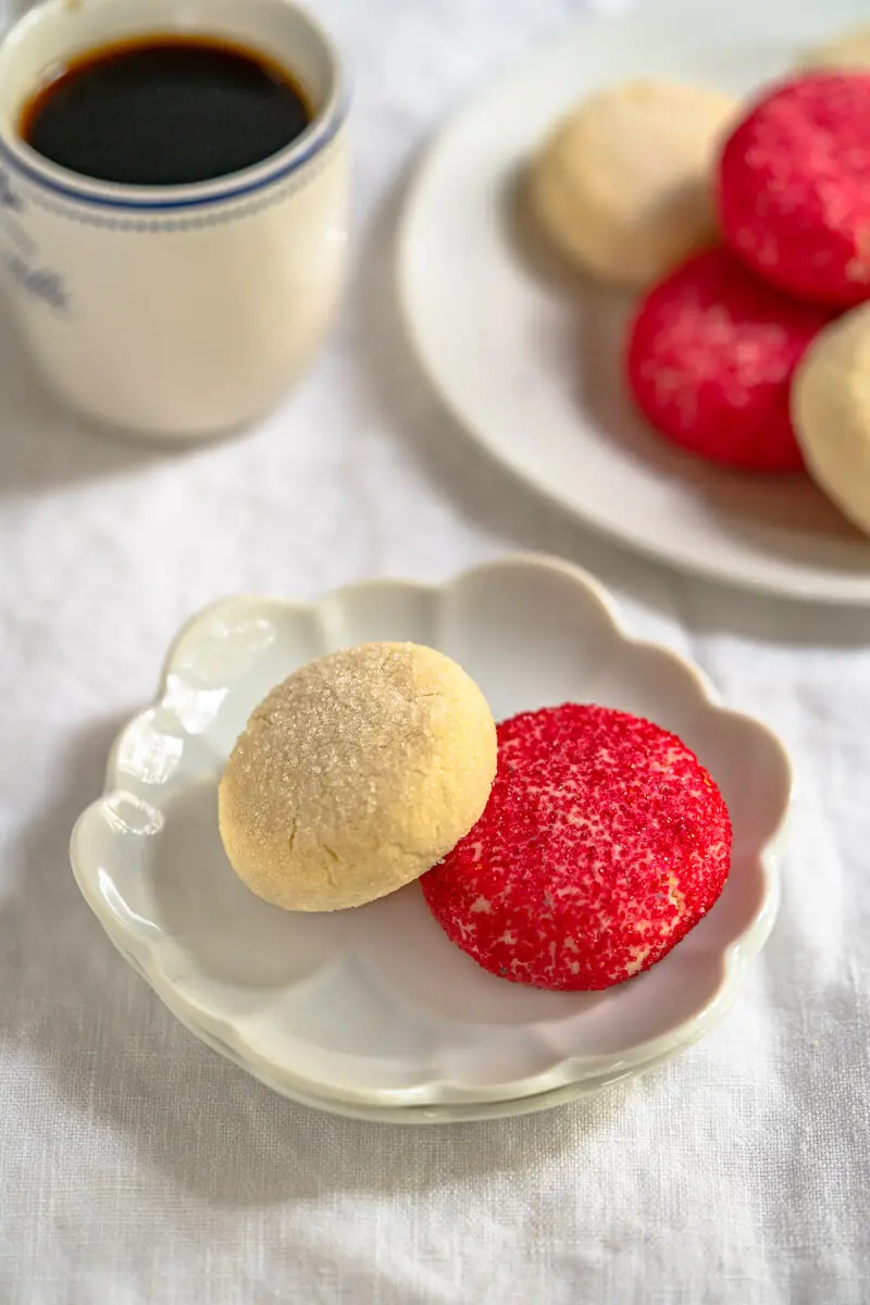 Angled view of a small white dish holding a red and a white decorated butter cookie with a cup of coffee and a plate of cookies in the background.