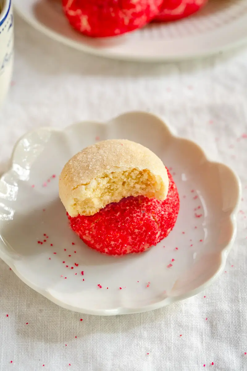 Side view of a white dish holding butter cookie decorated with red sprinkles with a butter cookie decorated with white sugar sitting on top of it with a bite out of it. A plate of cookies sit in the background.