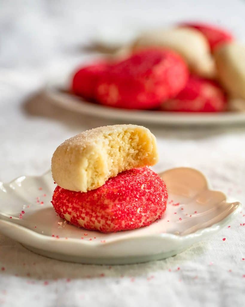 Side view of a white dish holding butter cookie decorated with red sprinkles with a butter cookie decorated with white sugar sitting on top of it with a bite out of it. A plate of cookies sit in the background.