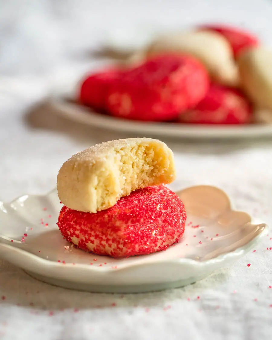 Side view of a white dish holding butter cookie decorated with red sprinkles with a butter cookie decorated with white sugar sitting on top of it with a bite out of it. A plate of cookies sit in the background.