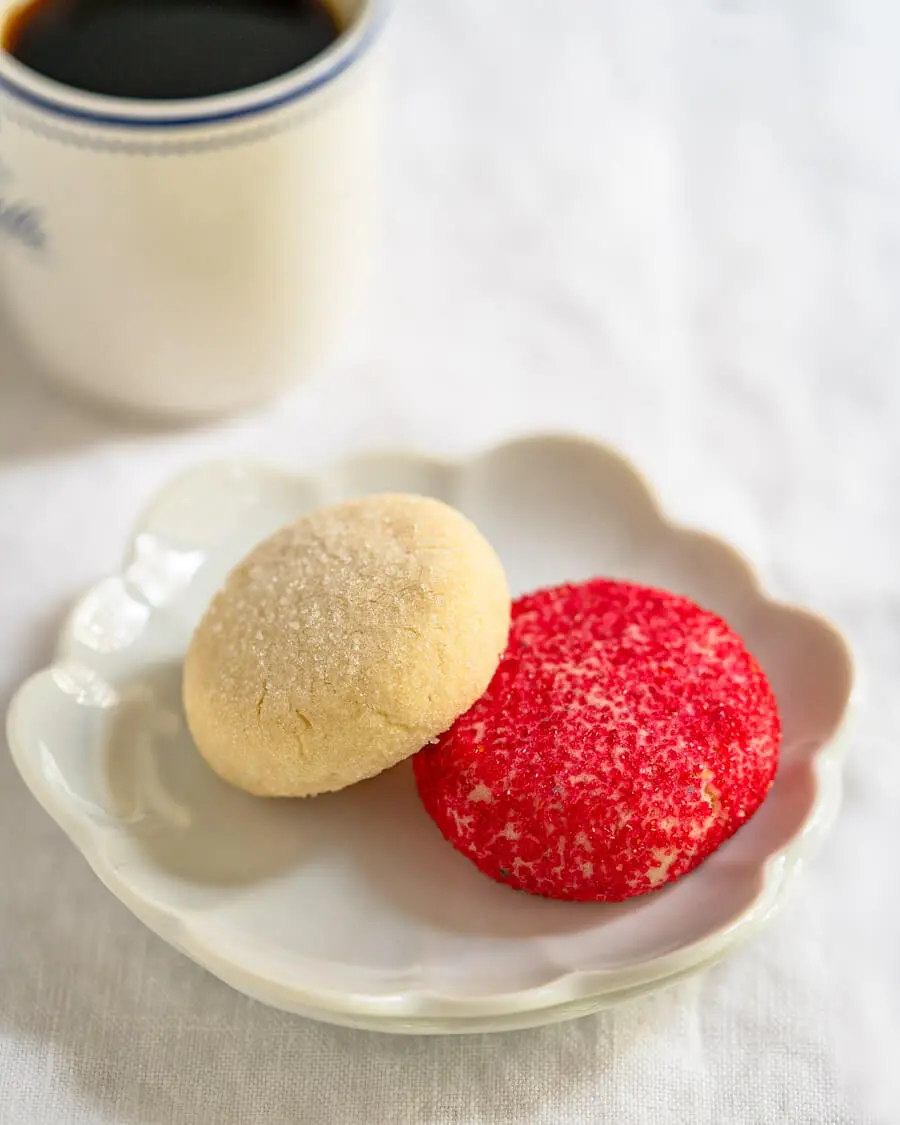 Angled view of a small white dish holding a red and a white decorated butter cookie with a cup of coffee in the background.