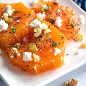 White plate with sliced oranges drizzled with a poppyseed vinaigrette and garnished with feta, walnuts, and sliced green onions.