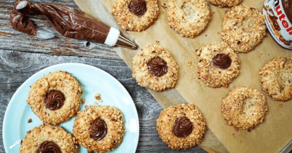 A top-down view of cookies on a plate and on a wooden cutting board. A piping bag full of Nutella and the jar of Nutella sit to the side.