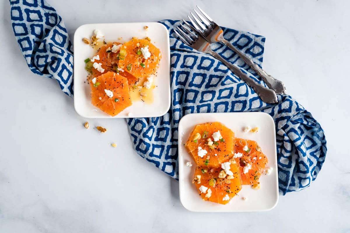 Top-down shot of two plates of fresh orange slices drizzled in a poppyseed vinaigrette and garnished with feta, sliced green onions, feta, and chopped walnuts.