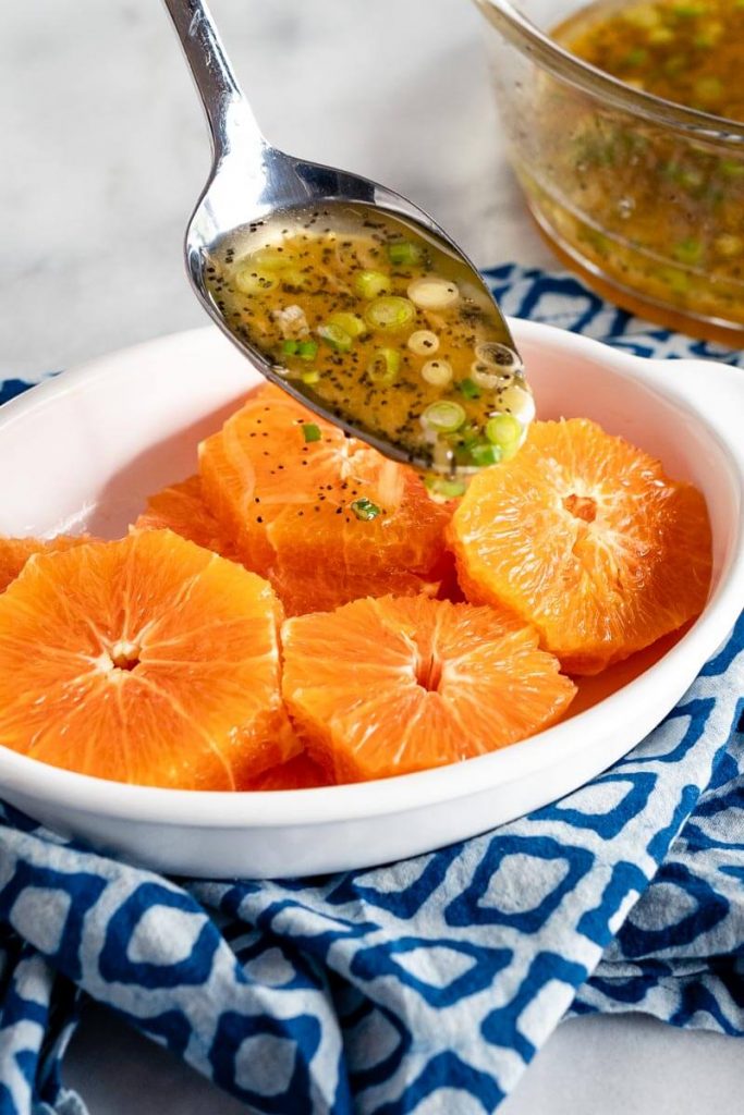 White oval dish filled with slices of fresh oranges with a spoon of poppyseed vinaigrette hovering over the oranges.