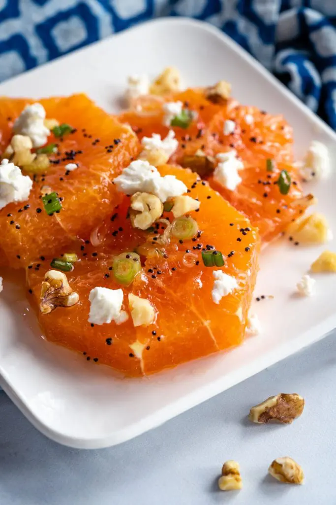 White dish filled with sliced oranges drizzled with a poppyseed vinaigrette and garnished with feta, walnuts, and sliced green onions.