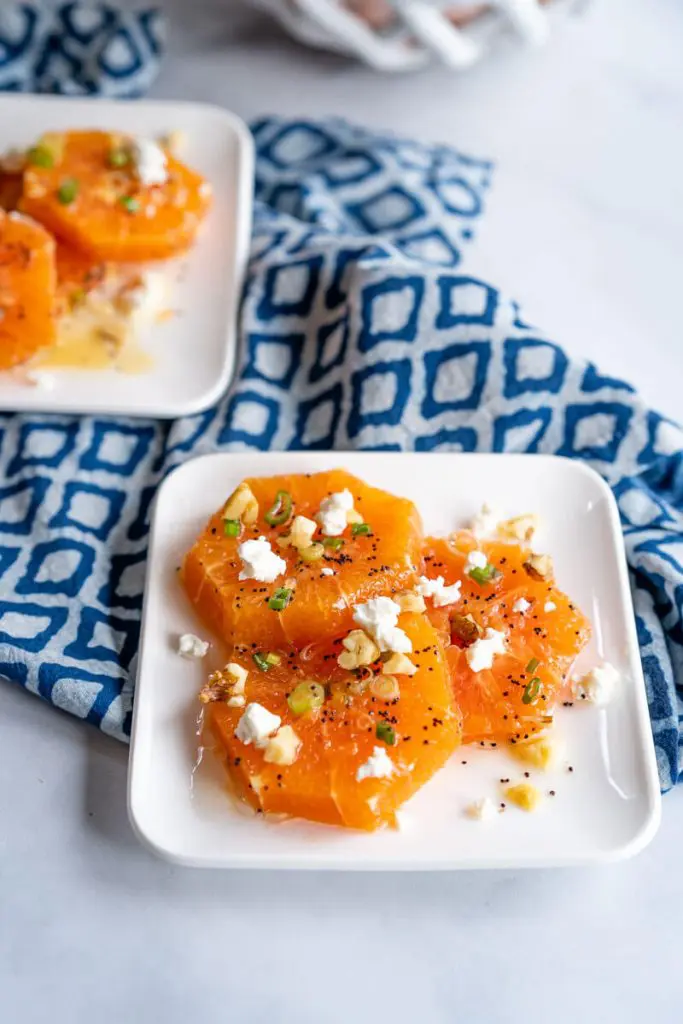 Top-down shot of two plates of fresh orange slices drizzled in a poppyseed vinaigrette and garnished with feta, sliced green onions, feta, and chopped walnuts.