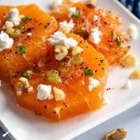 Close-up angle shot of a white plate holding fresh orange slices drizzled in a poppyseed vinaigrette and garnished with feta, sliced green onions, feta, and chopped walnuts.