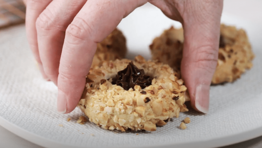 Sideview of three Nutella Filled Thumbprint cookies on a white plate. A hand is picking up the front one.