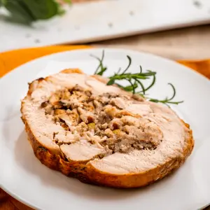 angled photo of a slice of turkey roulade stuffed with a swirl of stuffing on a white plate next to a rosemary garnish.