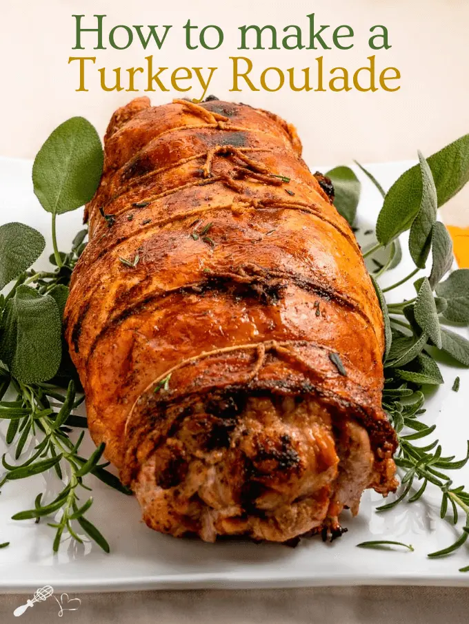 A front view of an uncut turkey roll tied with twine and cooled to a golden brown on a white platter garnished with fresh sage and rosemary.