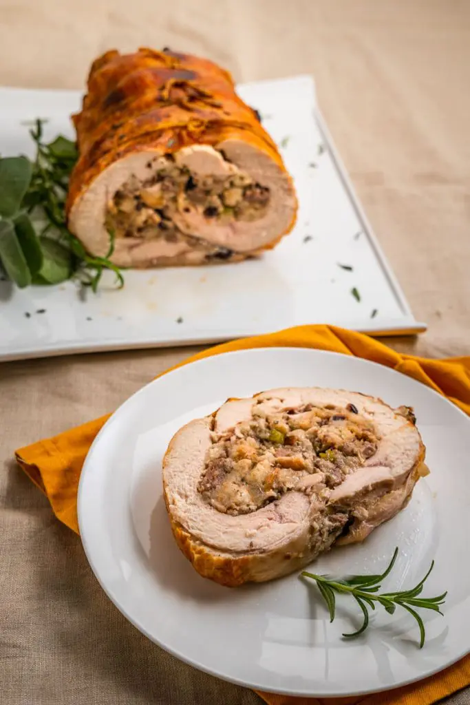 Top down view of a slice of turkey roulade swirled with dressing on a white plate. The whole turkey roll it the first slices removed is in the background.