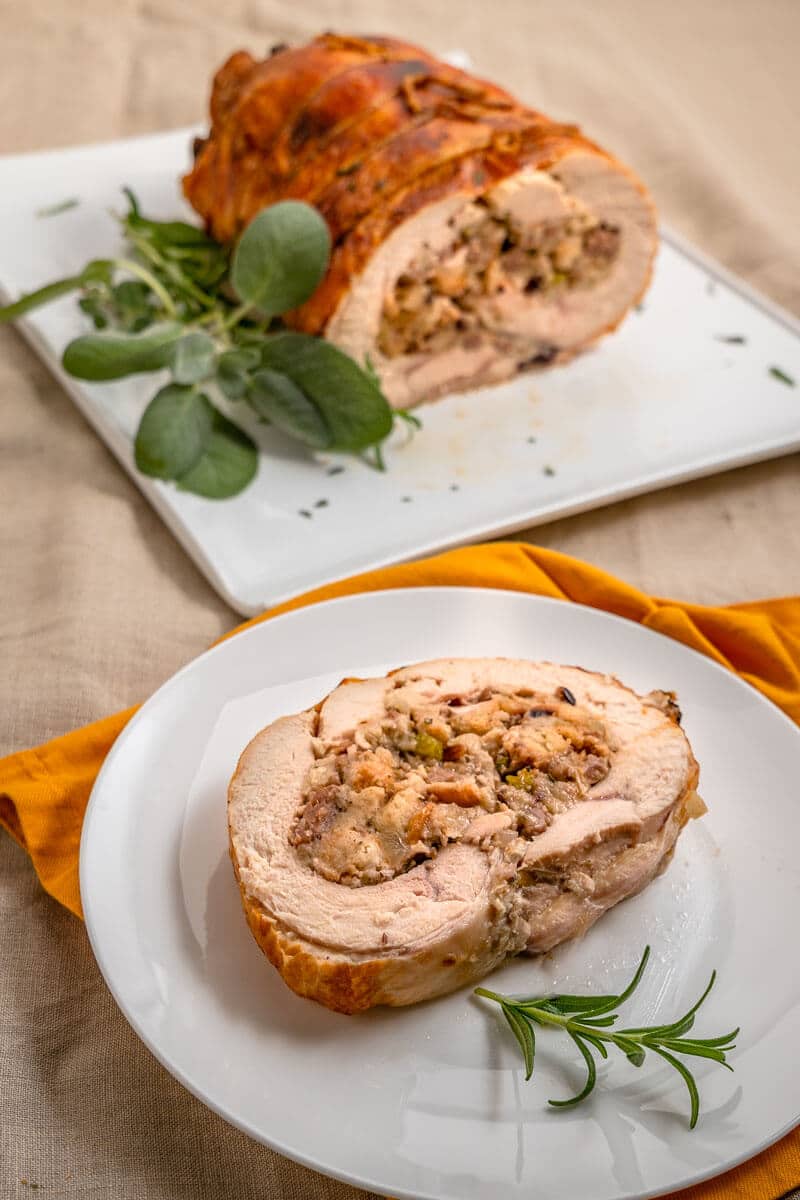 Top down view of a slice of turkey roulade swirled with dressing on a white plate. The whole turkey roll it the first slices removed is in the background.