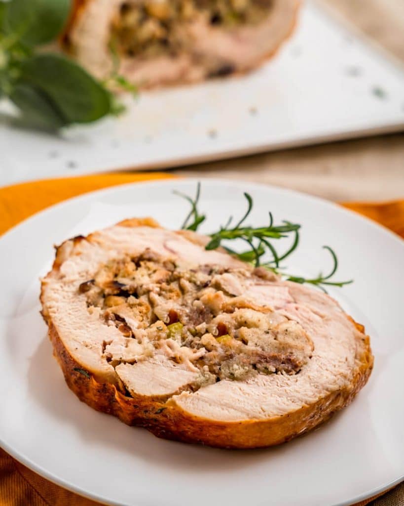 Top down view of a slice of turkey roulade swirled with dressing on a white plate. The whole turkey roll it the first slices removed is in the background.