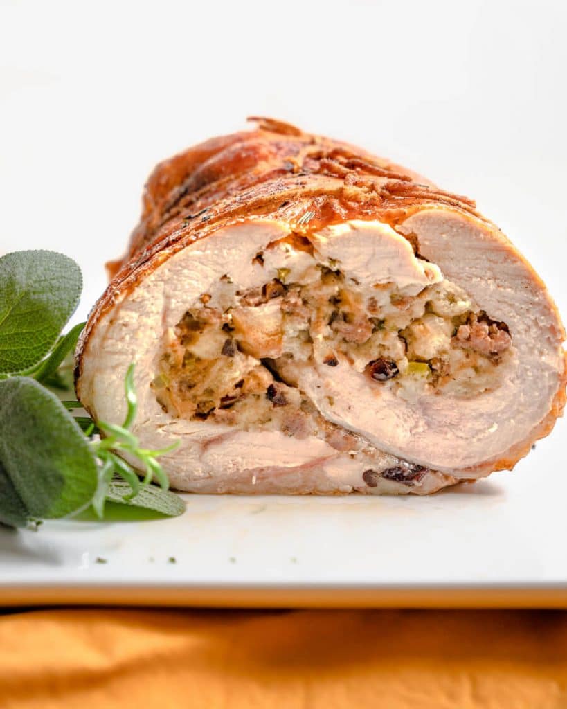 Front view of a sliced turkey roulade on a white platter garnished with sage and rosemary.