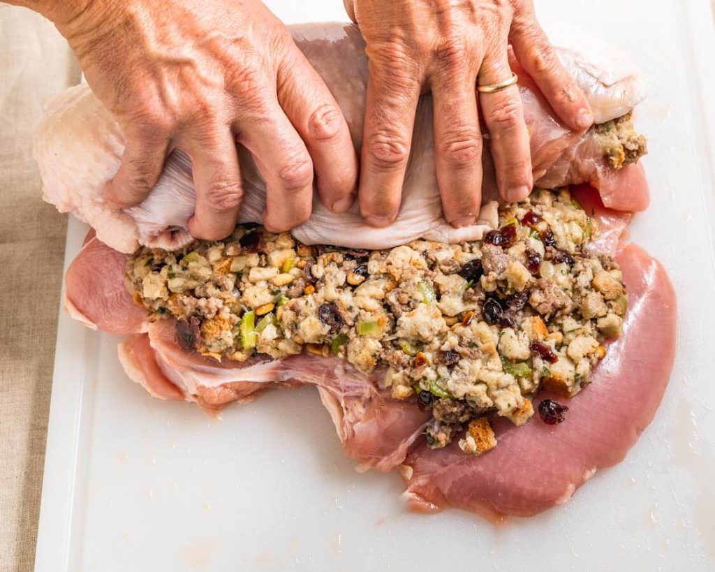 A butterflied turkey breast topped with stuffing being rolled tightly into a turkey roll.