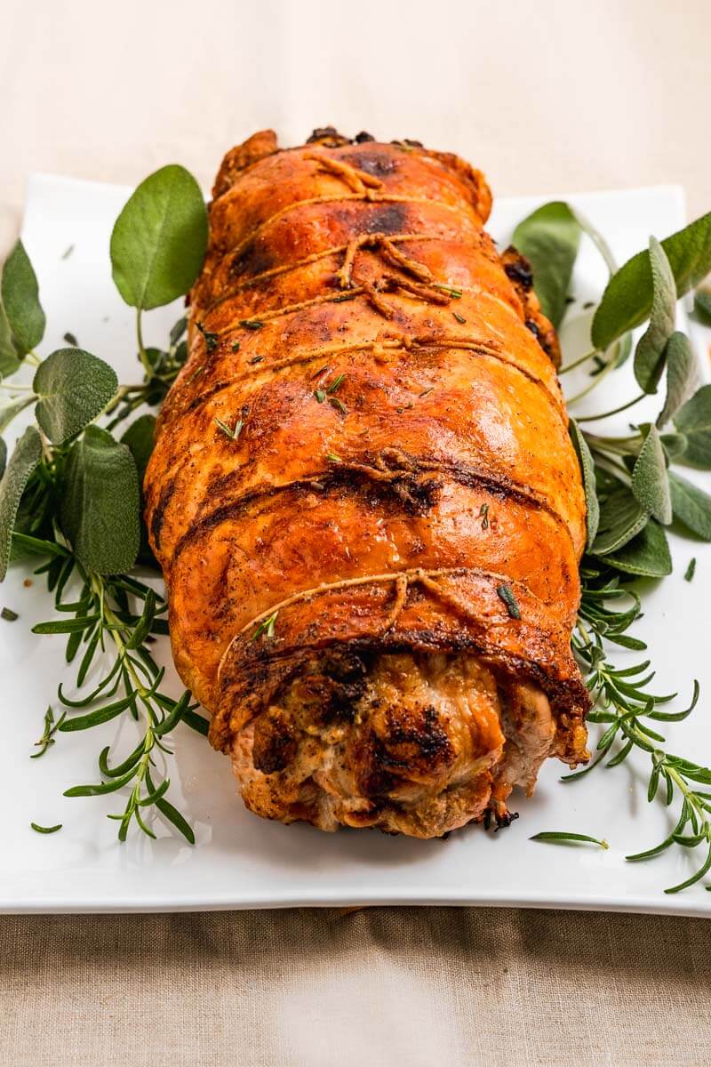 Top angled view of a turkey roulade tied and baked to a golden brown on a white platter and garnished with fresh sage and rosemary.