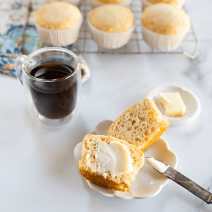 Top down photo of a muffin cut in half on a white plate with an antique knife propped against the plate next to a cup of coffee and a pat of butter. A cooling rack full of muffins sit in the background.
