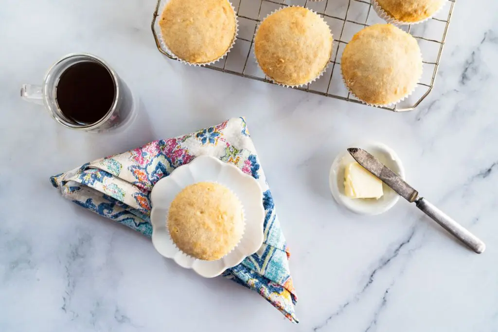 Top down photo of a muffin on a white plate over a multi-colored napkin next to a cup of coffee and a pat of butter. A cooling rack full of muffins sit in the background.