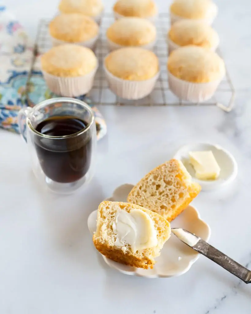 Top down photo of a muffin cut in half on a white plate over a multi-colored napkin next to a cup of coffee and a pat of butter. A cooling rack full of muffins sit in the background.