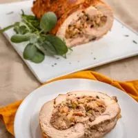 Top down view of a slice of turkey roulade swirled with dressing on a white plate. The whole turkey roll it the first slices removed is in the background.