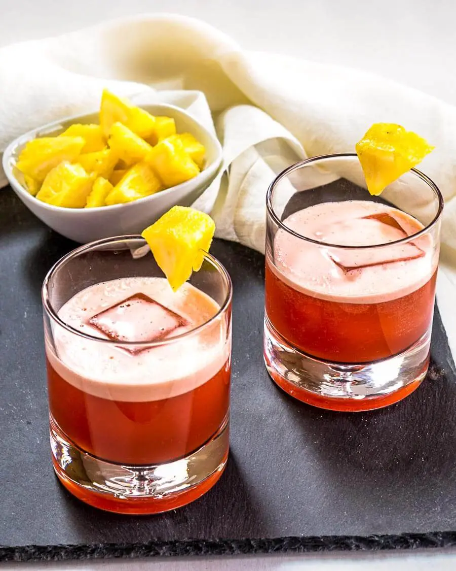 3/4 angle of two rocks glasses of a red Campari Cocktail garnished with pineapple sitting on slate with a bowl of pineapple in the background.