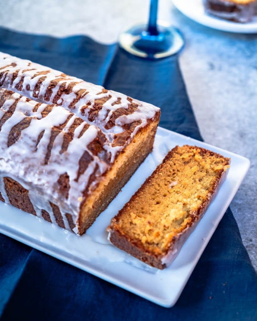 Top down view of a loaf of cake drizzled with sugar glaze sitting on a white platter. The front slice is sitting to the front of the loaf. The platter sits on a blue napking.