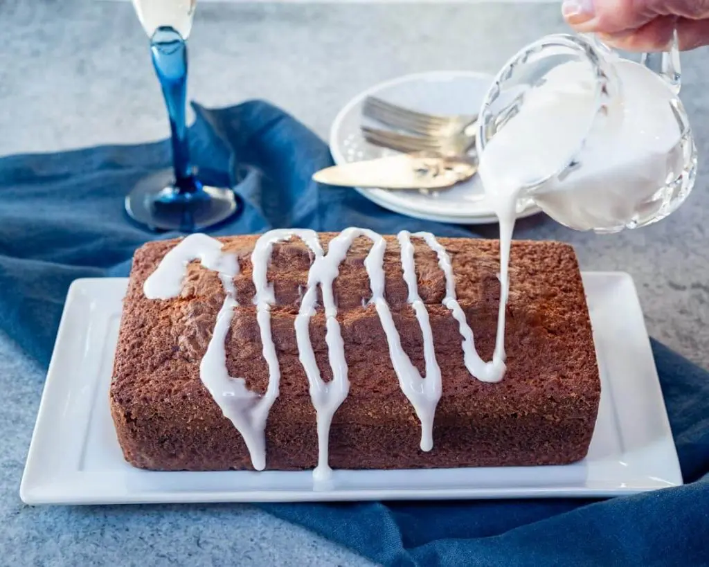 A loaf of champagne cake with a sugar glaze drizzling over the top sitting on a white plate over a blue napkin.