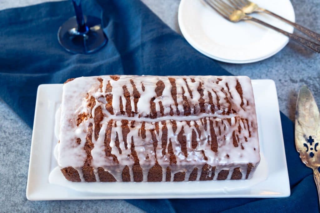 Top down view of a loaf of Champagne cake covered with a champagne drizzle sitting on a white platter over a blue napkin. A set of plates and a champagne glass sit int he background.