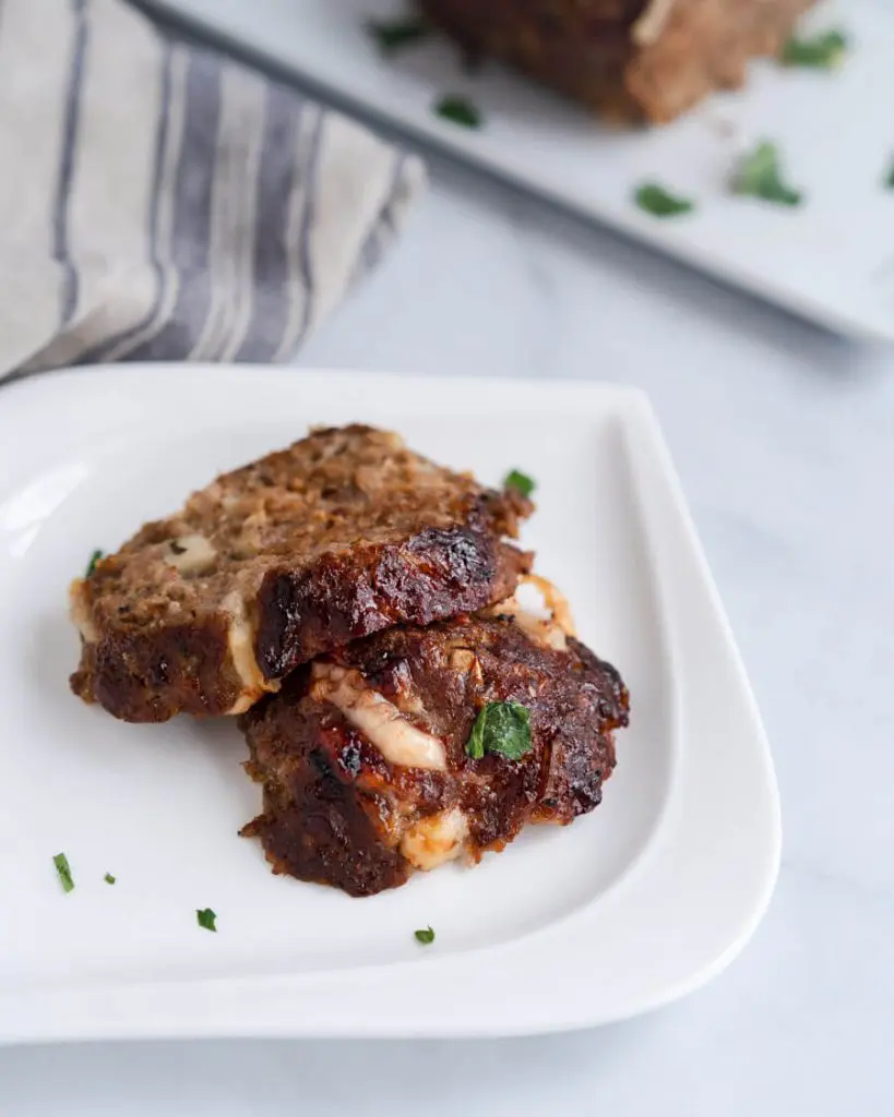 Top down photo of two slices of Cheese Stuffed Meatloaf sitting on a white plate. The plate is garnished with fresh parsley and sits on a blue stripped napkin.