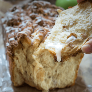 A hand holding a piece of cinnamon apple bread thats been pulled from the loaf. A drizzle of glaze is running off the front.