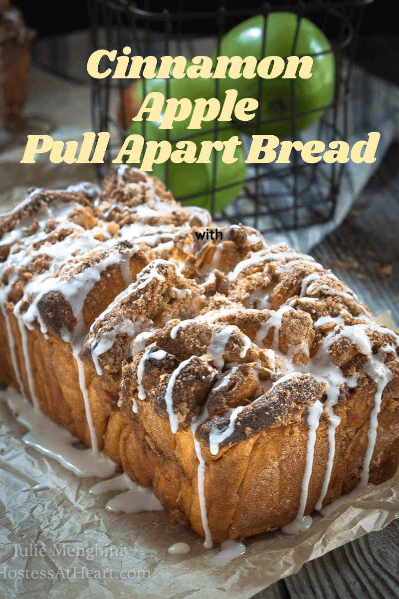 Sideview of a loaf of cinnamon apple pull apart bread topped with streusel and a drizzle glaze sitting on a piece of parchment paper and a basket of green apples sit in the background.
