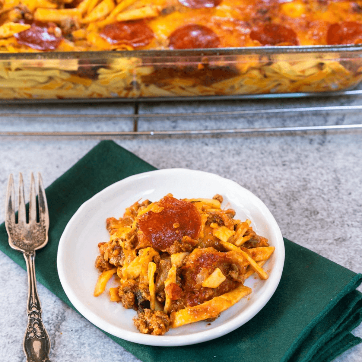 Top down angle view of a plate of pizza casserole topped with pepperoni on a white plate over a green napkin. A pan of the casserole sits in the back.