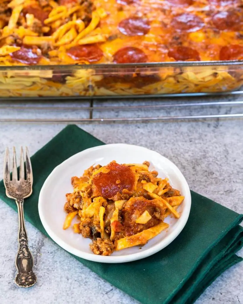 Top down angle view of a plate of pizza casserole topped with pepperoni on a white plate over a green napkin. A pan of the casserole sits in the back.