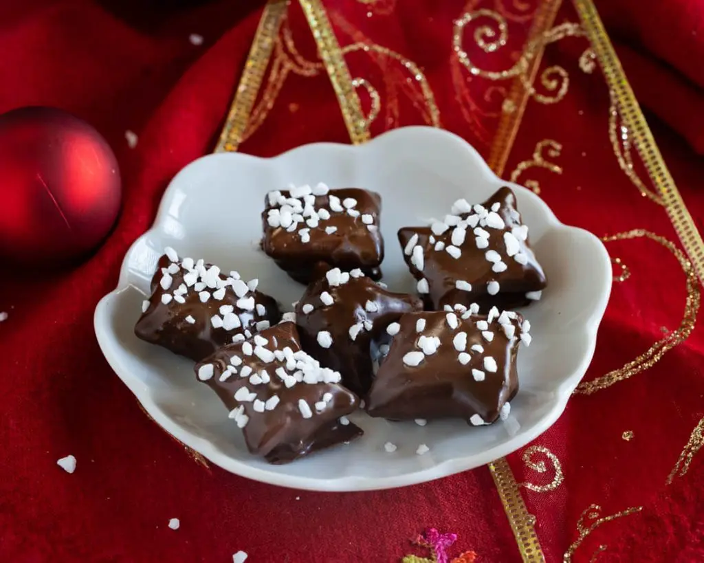 Top down view of chocolate covered Reindeer Food pretzel bites garnished with pearl sugar sitting on a white plate over a red napkin net to Christmas bulbs. A second plate sits in the background.