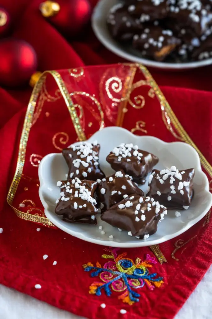 Close up sideview of chocolate covered pretzels Reindeer Snack bites topped with Swedish Pearl Sugar sitting on a white plate. A second bowl wits in the background.