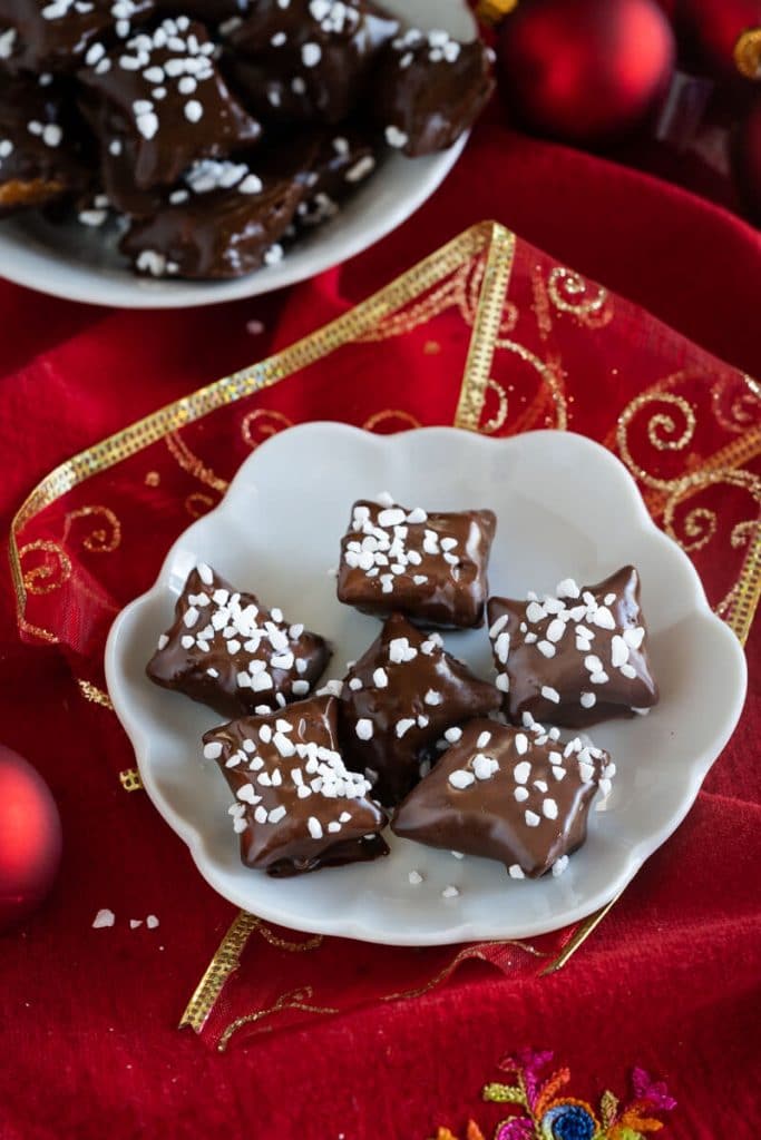 Top down view of chocolate covered pretzel bites garnished with pearl sugar sitting on a white plate over a red napkin net to Christmas bulbs. A second plate sits in the background.