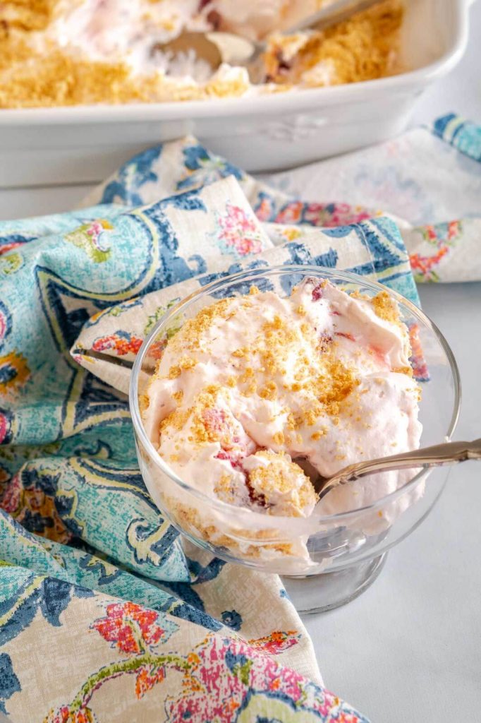 Top down view of a glass dish filled with a Strawberry Cheesecake Salad that's been garnished with graham cracker crumbs. A white baking dish filled with the salad sits in the background.