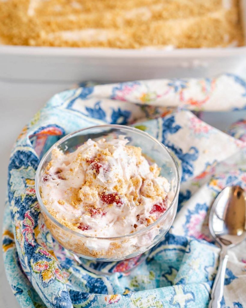 Top down view of a glass dish filled with a Strawberry Cheesecake Salad that's been garnished with graham cracker crumbs. A white baking dish filled with the salad sits in the background.